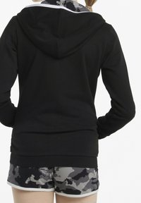 Persona con sudadera negra y pantalones cortos de camuflaje gris-negro, vista desde atrás sobre un fondo blanco liso.
