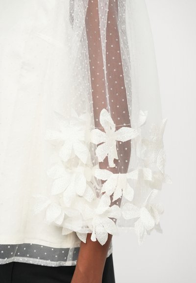 Bela bluzica s prozornimi rukavima s točkastim uzorkom i teksturiranim cvjetnim aplikacijama na manžetama. Ima laganu, prozračnu tkaninu s izraženim dizajnom.