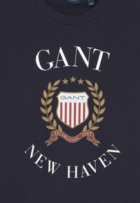 Námořnické tričko má logo GANT, erb s červenými a bílými pruhy, obklopený olivovými ratolestmi a textem "New Haven" a "Established 1949."
