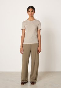 Young woman with curly hair wears beige short-sleeve shirt, loose olive-green pants, and brown shoes, standing against plain white background.