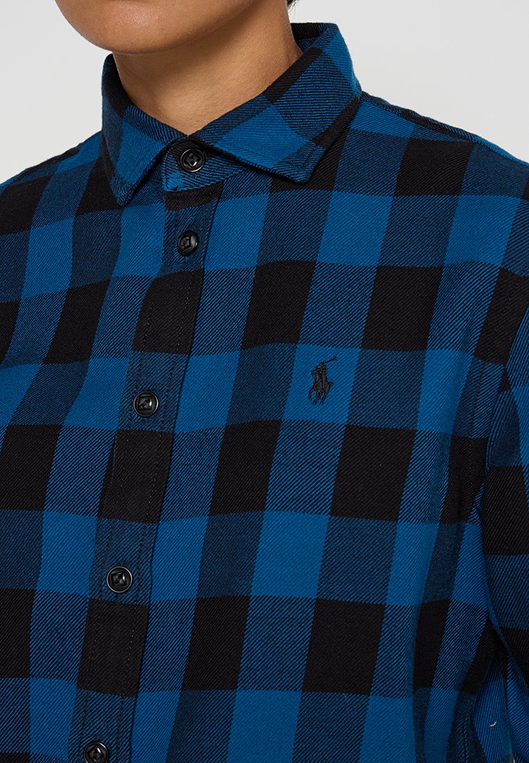 Camicia flanella a quadri blu e nera con colletto a punta, chiusura con bottoni e piccolo logo sul petto. Tessuto strutturato con vestibilità regolare.