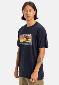 Camiseta de algodón color azul marino con un estampado gráfico en la parte delantera que presenta un atardecer, palmeras y olas. Cuello redondo y mangas cortas.