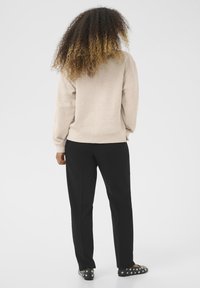 Bežová mikina s voľným strihom, čierne nohavice a čierne slip-on topánky zdobené striebornými cvočkami. Textúrované vlasy padajú cez chrbát.