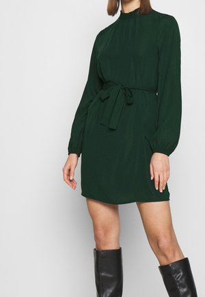 Kvinde iført en mørkegrøn minikjole med lange ærmer og et bindebælte i taljen samt sorte knæhøje støvler, stående mod en ensfarvet baggrund.