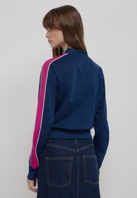 Giacca da track blu navy con colletto alto, con strisce laterali rosa e bianche, e un tessuto liscio e elasticizzato. Indossata con una gonna di denim scuro.