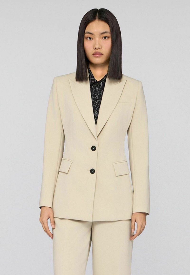 Beige op maat gemaakte blazer met een single-breasted ontwerp, voorzien van twee for pocketen en zwarte knopen, gedragen over een zwart patroonhemd.