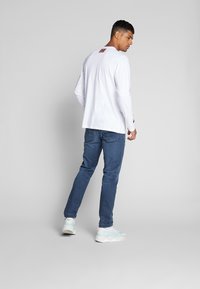 Cămașă albă cu mâneci lungi, cu logo pe spate, asortată cu blugi albaștri și adidași deschiși, evidențiind un fit casual și un design curat.