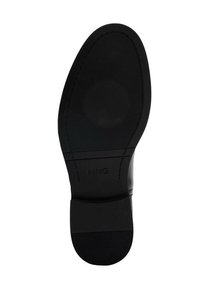 Suela de zapato negra hecha de goma con una superficie texturizada. Presenta un diseño circular y la marca "MNG," con una forma redondeada y ranuras visibles.