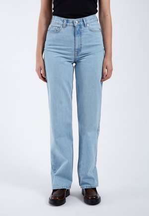 Personne portant un jean bleu clair taille haute à jambes droites, un haut noir et des chaussures marron foncé, debout sur un fond blanc uni.