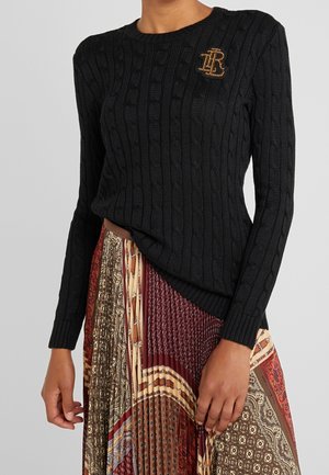 Czarny sweter w splot warkocza z złotą broszką z monogramem na piersi, zestawiony z wielokolorową plisowaną spódnicą ozdobioną misternymi wzorami.