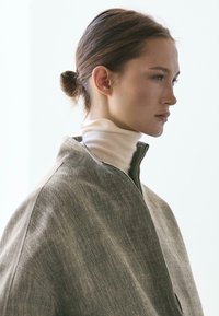 Giovane donna con i capelli raccolti in uno chignon, indossa un dolcevita crema a collo alto e una giacca grigia testurizzata con cerniera, rivolta verso destra su uno sfondo chiaro.