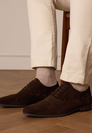 Chaussures habillées en daim marron avec un toe pointu, des coutures détaillées et des lacets. Associées à des chaussettes claires à côtes et à un pantalon beige.