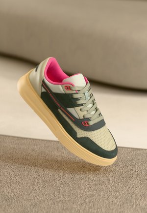 Zapatilla casual con detalles en beige, verde oscuro y rosa, suela beige y cordones grises, sobre una superficie de alfombra texturizada.