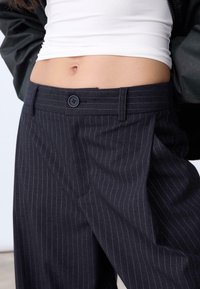Des pantalons rayés bleu marine foncé présentent un design taille haute, une fermeture à bouton sur le devant et un tissu doux et texturé avec une coupe ample.
