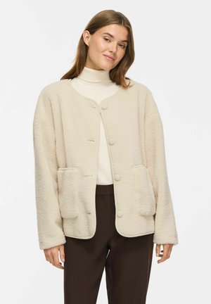 Femme portant un cardigan doux couleur crème avec de grandes poches devant, un col roulé blanc et un pantalon marron foncé sur un fond uni.
