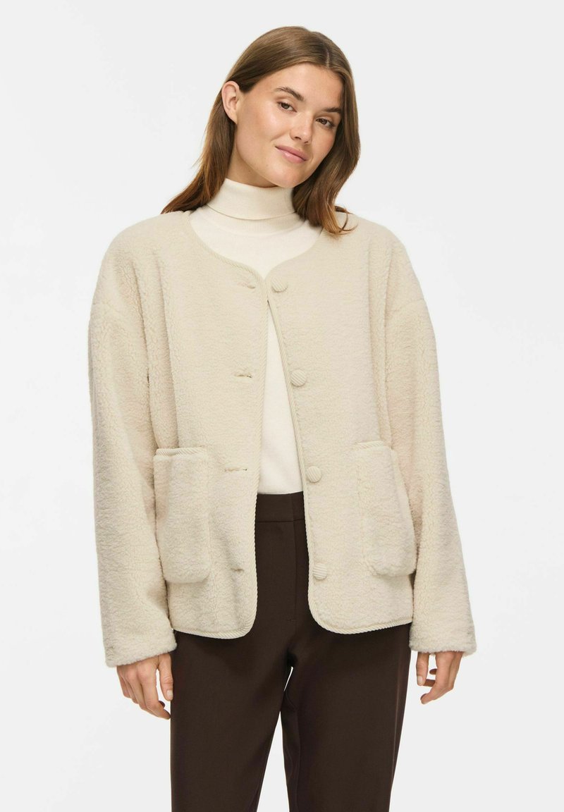 Femme portant un cardigan doux couleur crème avec de grandes poches devant, un col roulé blanc et un pantalon marron foncé sur un fond uni.