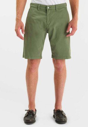 Olijfgroene casual herenshorts gedragen met zwarte veter-mocassins en een wit topje, zichtbaar van taille tot voeten.
