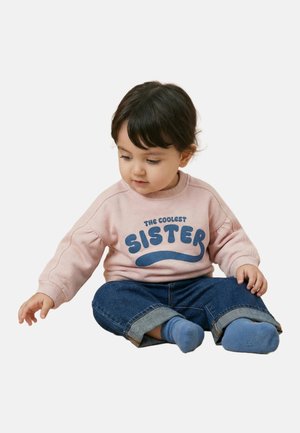 Un tout-petit aux cheveux foncés est assis les jambes croisées, portant un sweat-shirt rose avec l'inscription "The Coolest Sister", un jean bleu et des chaussettes bleues.