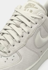 Sapatilhas Nike Air de tom cinza claro, com parte superior em camurça macia, logo em couro suave e atacadores planos. Apresenta perfurações na ponta e um colarinho acolchoado.