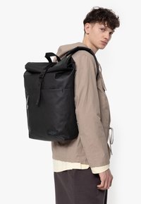 Sac à dos noir à rabat, doté d'une texture lisse et mate, de bretelles ajustables, d'une poche zippée à l'avant et d'un écusson logo en bas.