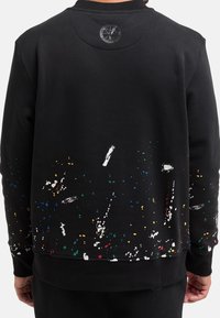 Černá mikina s texturovanou látkou; má barevný vzor cákanců barvy a kulatou nášivku s logem na krku.