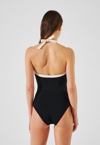 Costume da bagno nero con collo a halter e accento crema, dotato di una finitura testurizzata e un fiocco sul retro. Apertura della gamba alta.