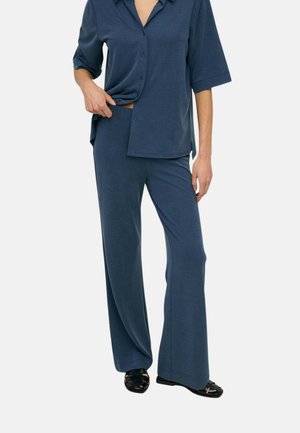 Mujer con blusa suelta azul oscuro de manga corta con botones y pantalones de pierna ancha a juego con zapatos negros de punta abierta.