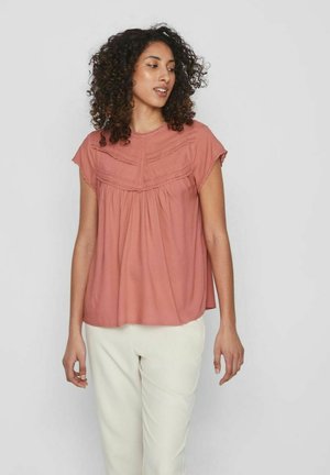 Hnedo-oranžová blúzka s krátkymi rukávmi a plisovanými detailmi na jarmu, vyrobená z ľahkej tkaniny a uvoľneného strihu, skombinovaná s krémovými nohavicami.