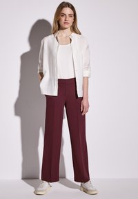 Chemise blanche à boutons portée par-dessus un débardeur blanc, assortie à un pantalon large bordeaux et des baskets blanches. Tissu lisse et coupe décontractée.