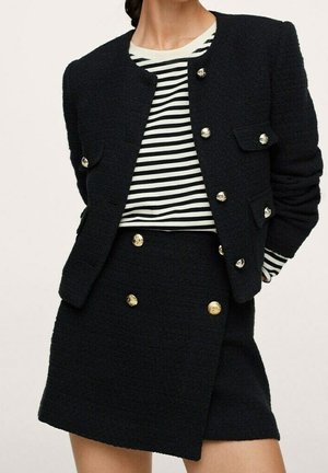 Femme portant un blazer texturé noir et une jupe avec des boutons dorés, associée à une chemise à manches longues noire et blanche à rayures.