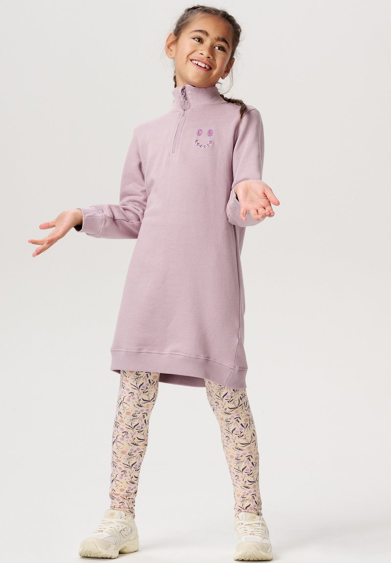 Lilla fleecekjole med høj krave og et smilende ansigtsdetalje, parret med mønstrede cremefarvede leggings. Hvide sportssko fuldender looket.