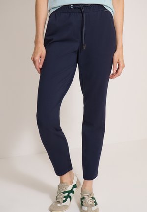 Femme portant un pantalon bleu marine à cordon de serrage et des baskets beige avec des rayures vertes, debout devant un fond clair uni.