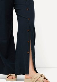 Pantalons larges de couleur marine avec des fentes sur les côtés, ornés de boutons marron le long de la longueur. Un tissu léger et texturé complète des sandales beige à nouer.