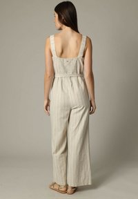 Combinaison beige rayée avec des jambes larges, un dos froncé et des bretelles réglables. Fabriquée en tissu léger pour plus de confort.