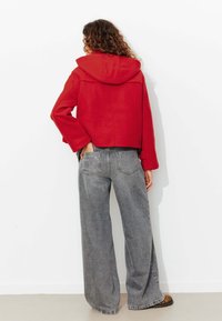 Veste rouge courte avec capuche, manches décontractées et détails de couture, associée à un jean gris à taille haute avec des jambes larges et des chaussures à imprimé animal.