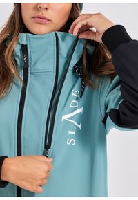 Teal jack met zwarte accenten, een volledige ritssluiting en een logo. Soepele textuur met drawstrings bij de kraag voor aanpassing.