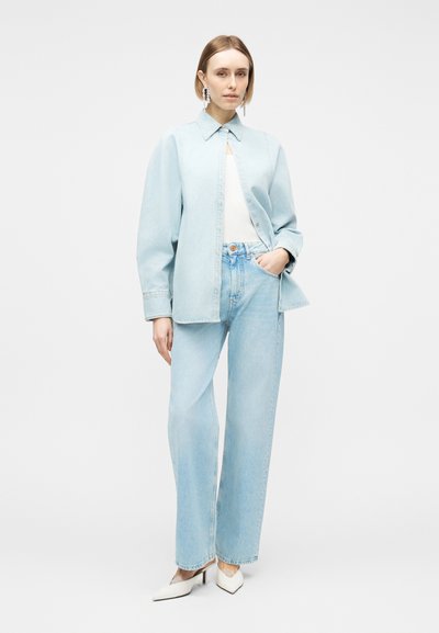 Femme debout portant une chemise en denim bleu clair, un jean large assorti, un haut blanc, des talons pointus blancs, avec des cheveux courts et de longues boucles d'oreilles.