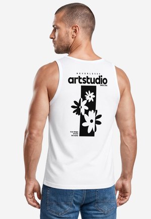 Man facing sideways wearing white sleeveless shirt with black floral design and text reading "artstudio" on the back, paired with blue jeans.