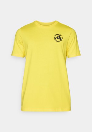Gul bomulds T-shirt med korte ærmer og rund halsudskæring. Har et sort cirkulært logo med tekst på venstre brystområde.