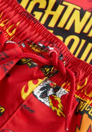 Punaiset uimashortsit, joissa on kiristysnyöri vyötäröllä, kiiltävä pinta sekä moniväriset liekkien ja sarjakuvahahmojen graafiset kuviot.