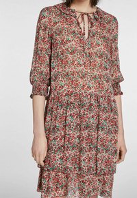 Robe florale en tissu léger et transparent. Présente des motifs rouges et verts, un col à nouer et des poignets élastiques. Ourlet évasé et en étages.