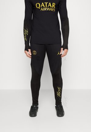 Mężczyzna ubrany w czarny strój sportowy z żółtym logo "Qatar Airways" na górze, napisem "Paris" i herbem klubu na spodniach, stojący na jasnoszarej podłodze.