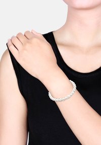 Femme en haut noir sans manches portant un bracelet blanc orné de perles à son poignet, une main reposant sur son épaule opposée.