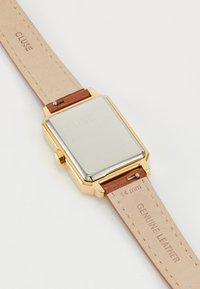 Montre rectangulaire dorée avec un dos en métal poli, bracelet en cuir véritable marron et détails en relief sur le bracelet.