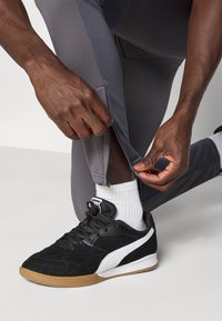 Μαύρο αθλητικό παπούτσι με λευκό λογότυπο, υφή στο επάνω μέρος και καουτσούκ σόλα· γκρι αθλητικό παντελόνι με φερμουάρ στον αστράγαλο· χέρια που ρυθμίζουν το παντελόνι.