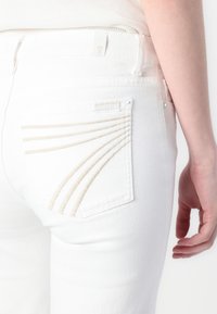 Jean en denim blanc avec une poche arrière ornée de rayures beiges texturées et d'une étiquette de marque, présentant un design ajusté et lisse.