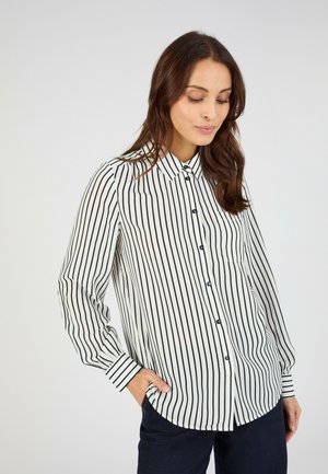 Femme aux longs cheveux bruns portant une chemise blanche à boutons avec des rayures verticales noires et un pantalon foncé, debout avec une main dans la poche.