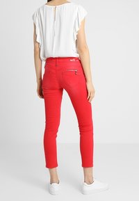 Jeans skinny rojos con una textura suave y una forma ajustada, combinados con una blusa blanca con volantes que presenta un detalle de ojal en la parte posterior.