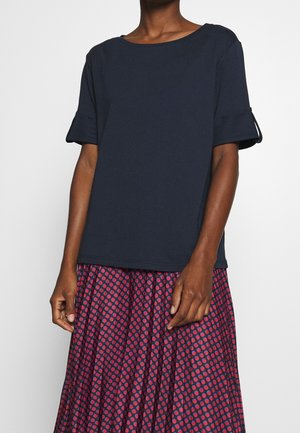 Kobieta w granatowej bluzce z krótkim rękawem z podwiniętymi mankietami i plisowanej czerwono-granatowej spódnicy w groszki, stojąca na jednolitym tle.