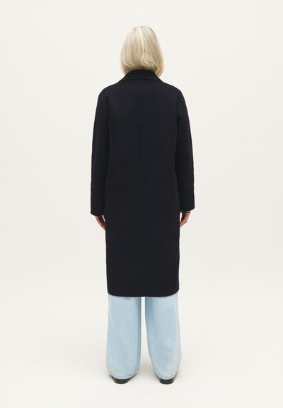 Cappotto lungo navy con taglio dritto e colletto classico, abbinato a pantaloni a gamba larga azzurri. Tessuto liscio, senza accessori visibili.